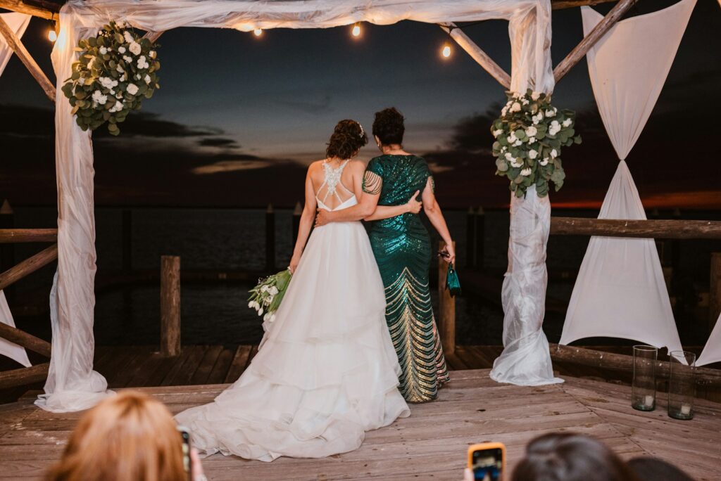 Two women embrace under a wedding arch at dusk.