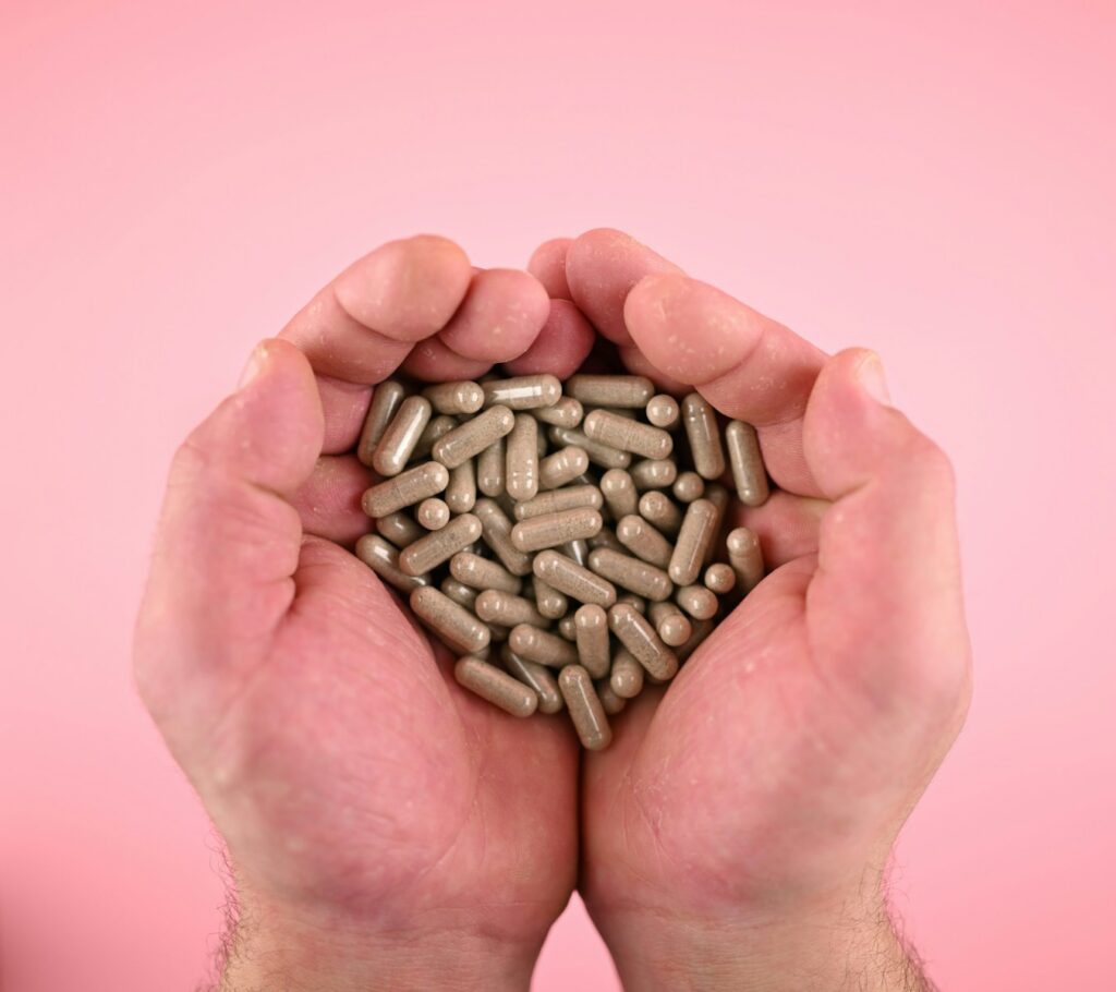 Hands holding numerous grey capsules on a pink background.