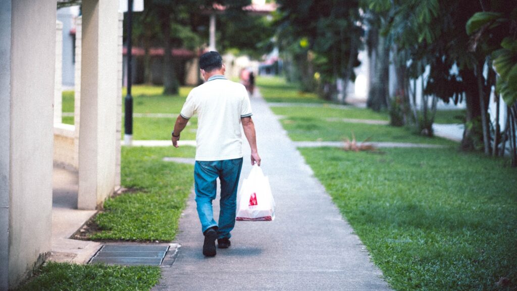 man in white t-shirt and blue denim jeans holding white plastic bag walking on sidewalk