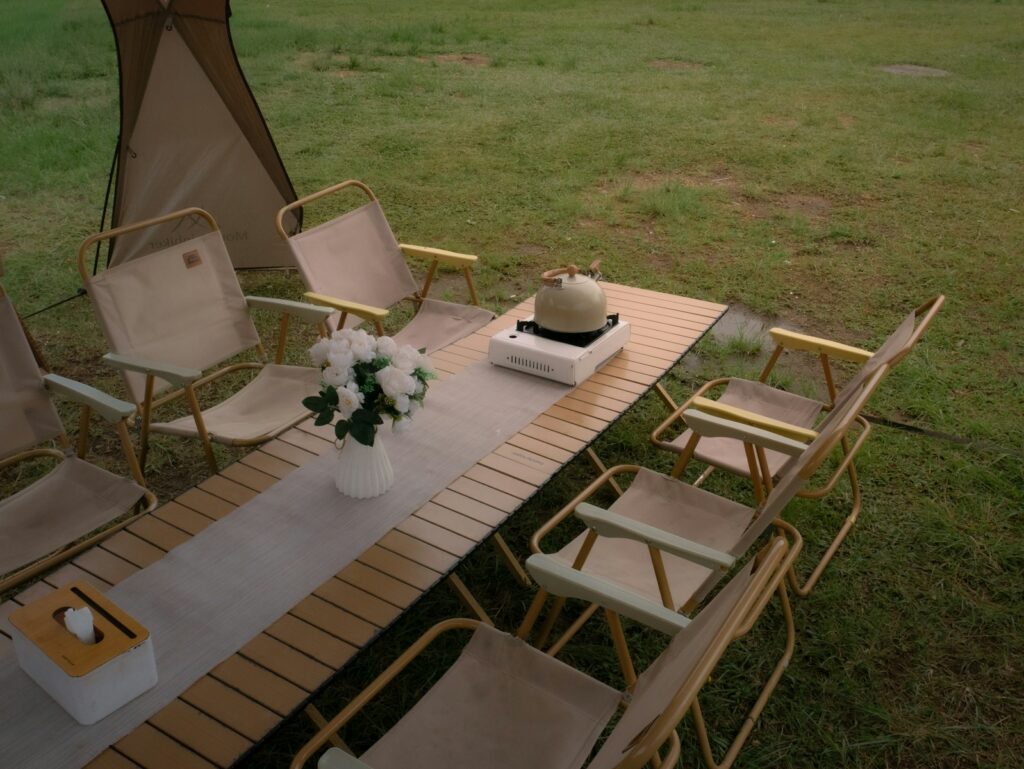 Outdoor camping table with chairs, flowers, and kettle on grass.