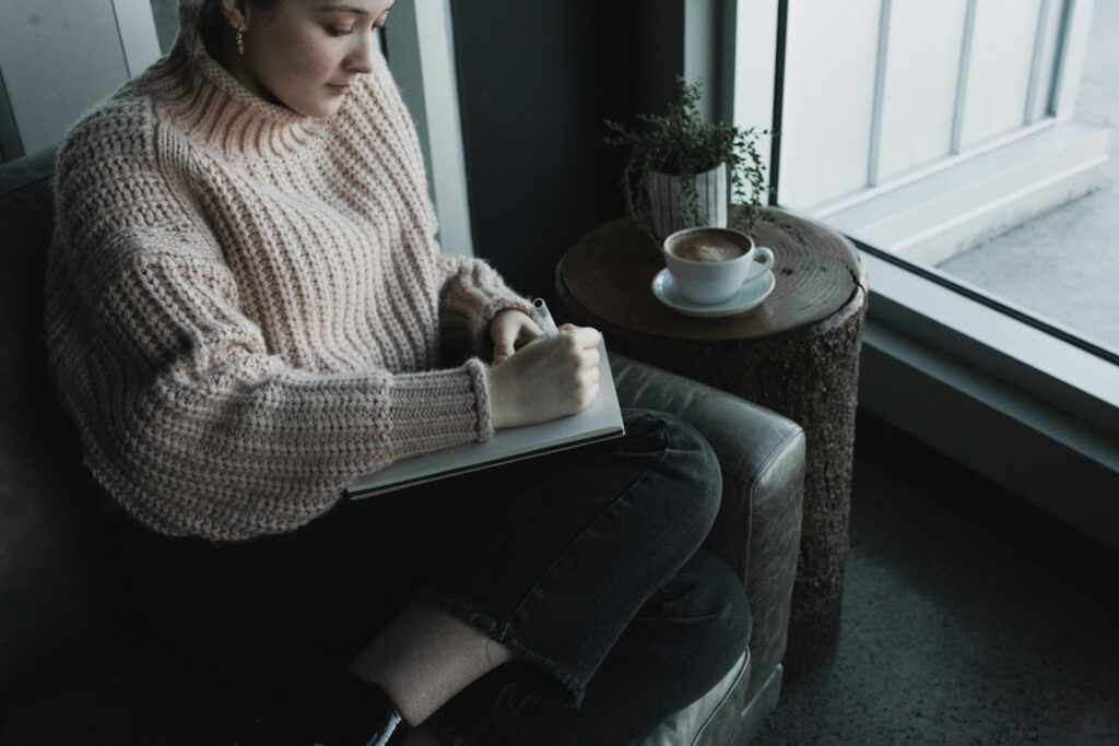 woman in white knit sweater and blue denim jeans writing morning intention in journal.