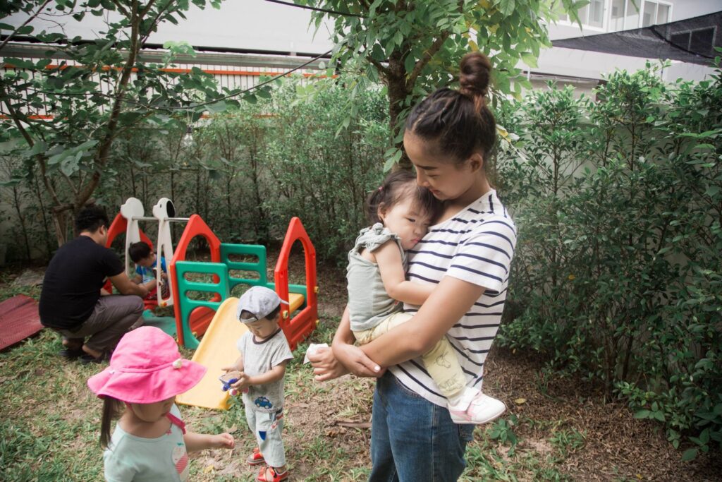 A serene backyard scene with a mother holding her child while kids play in the background.