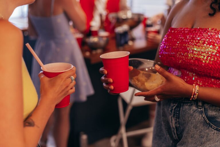 Two women holding red party cups and snacks at a casual indoor gathering.