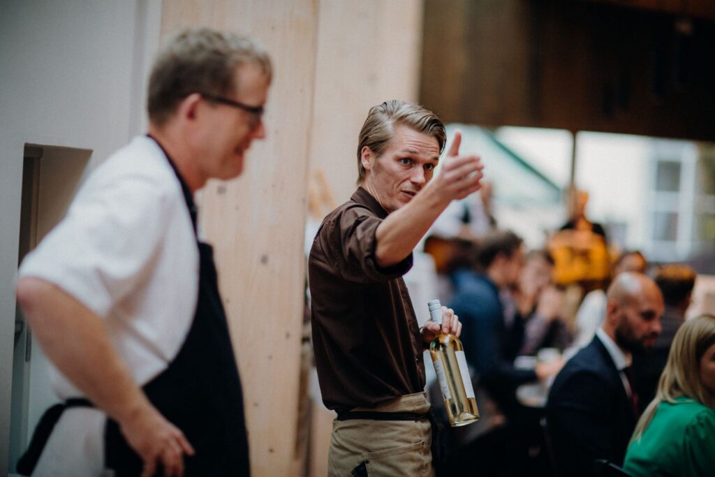 Waiter gesturing while holding wine bottles at a lively restaurant setting.