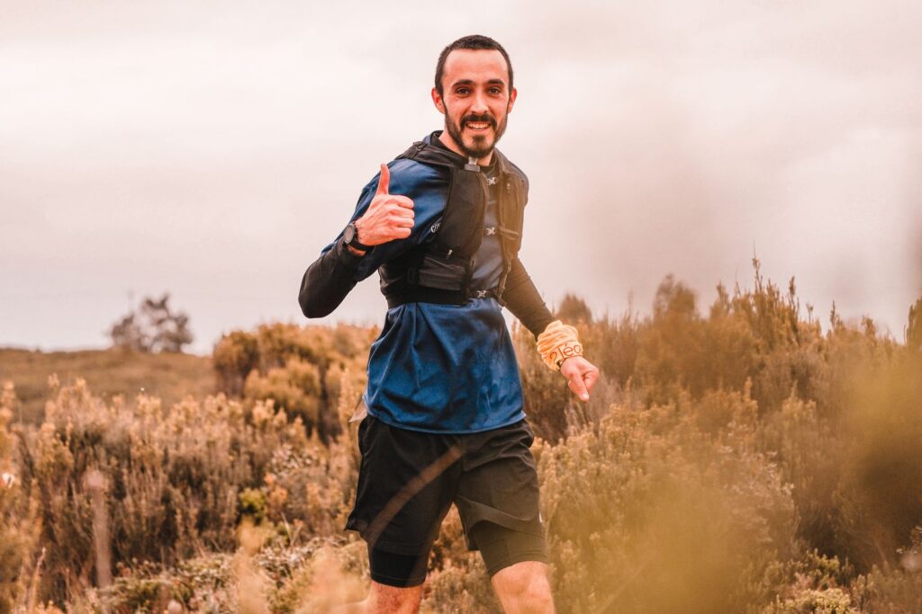 Smiling man running outdoors in nature with a thumbs up gesture, showcasing positivity and fitness.