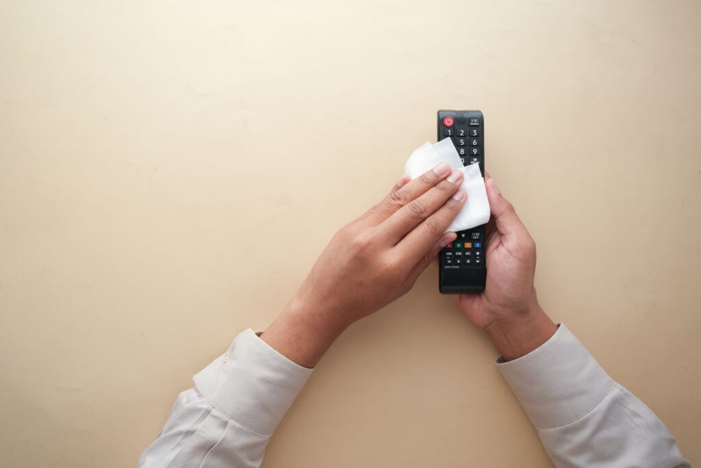 Hands cleaning a TV remote control with a cloth on a beige background for hygiene and cleanliness.