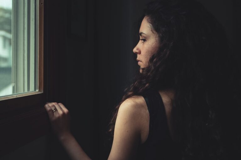 A brunette woman with long hair gazes thoughtfully out the window indoors.