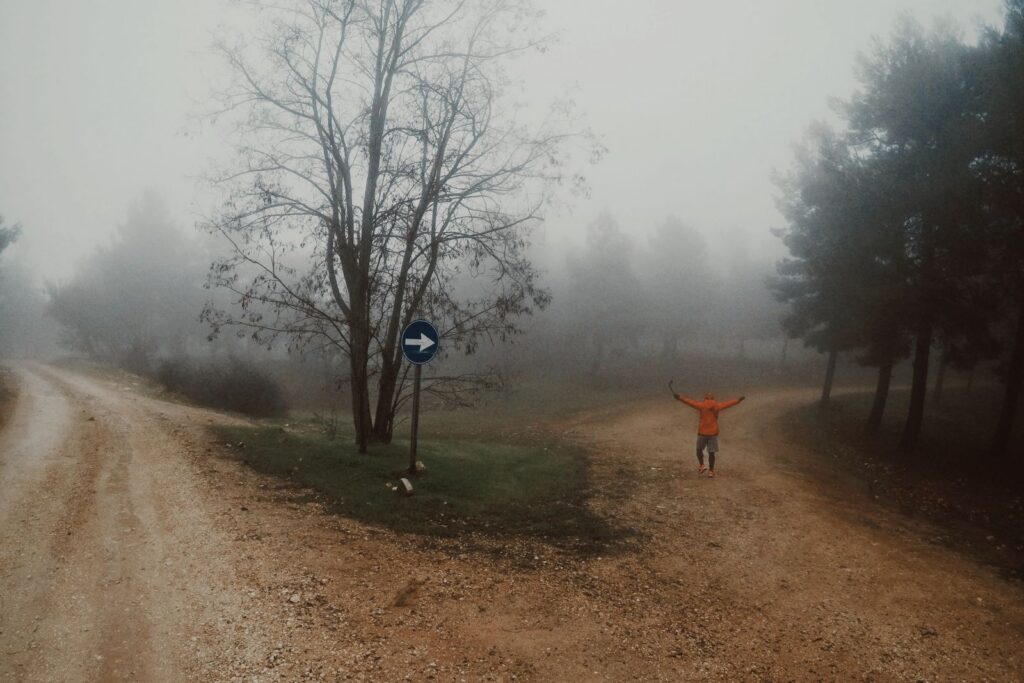 A person in an orange jacket explores misty dirt roads in a foggy forest setting.