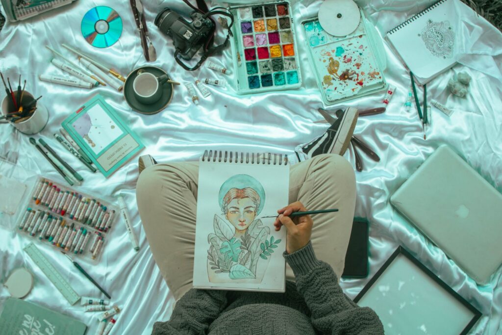 Top view of an artist's workspace with sketchbook, art supplies, and digital camera on a satin background.