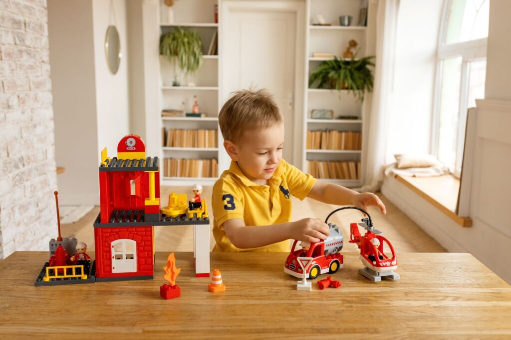 A young boy in a yellow shirt playing with toy building blocks and vehicles on a wooden table.