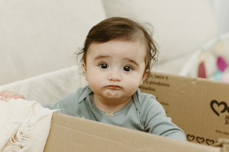 Cute baby sitting in a cardboard box with a confused expression, indoors.