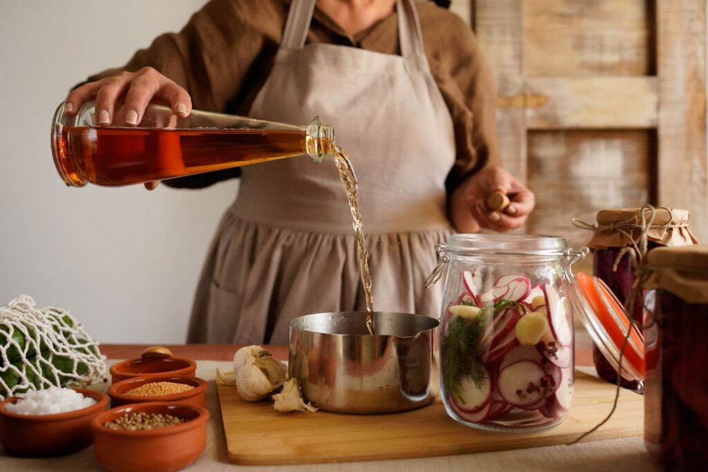 A woman pours vinegar into a pot while making homemade pickles in a rustic kitchen setting.