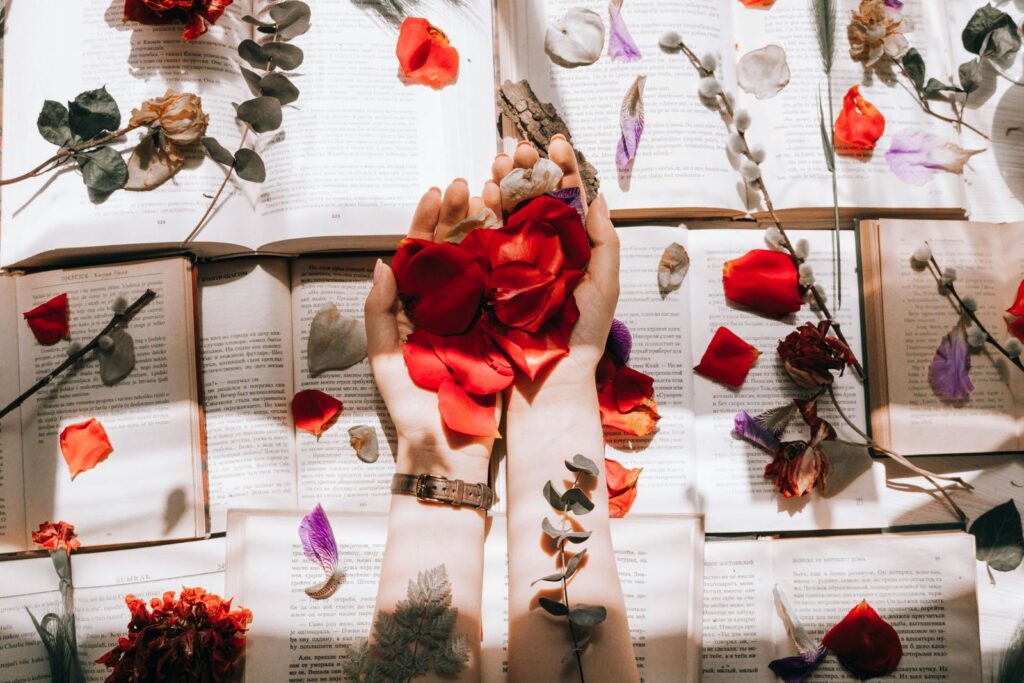 Hands with red petals over open books, surrounded by scattered flowers and petals for artistic composition.