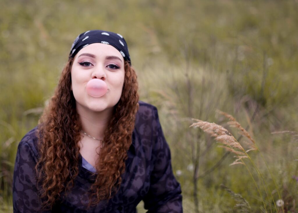 A cheerful young woman blows a bubble gum bubble while sitting in a field.