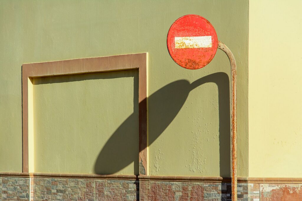 A weathered 'No Entry' sign casting a shadow on a textured wall, captured in bright daylight.