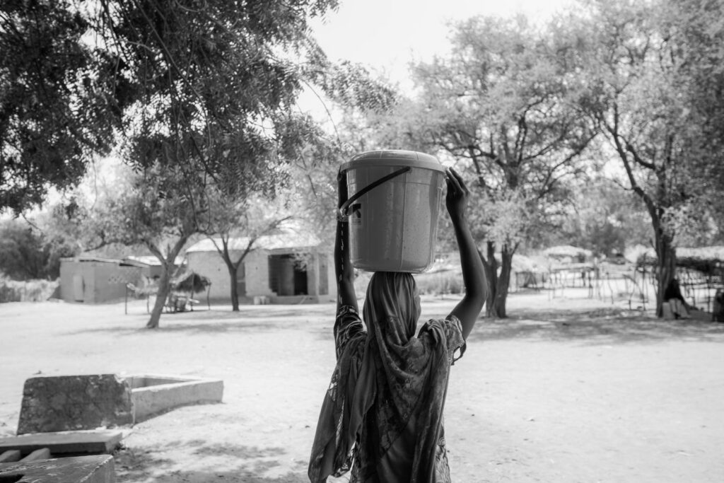 A woman carrying a water container on her head in a rural village environment.