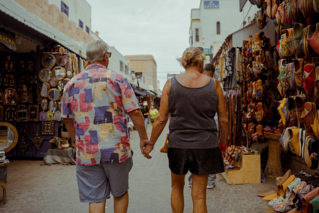 Senior couple holding hands in Essaouira's colorful street market, filled with Moroccan art and crafts.