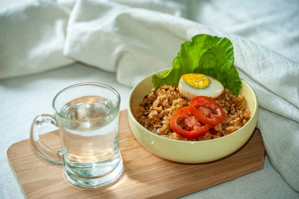 Tasteful bowl of fried rice with tomato, lettuce, and egg, served with water on a wooden board.