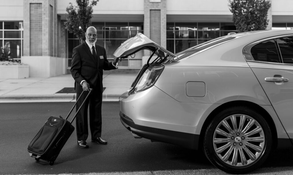 Chauffeur driver in suit loading luggage into luxury car trunk outside a modern building.
