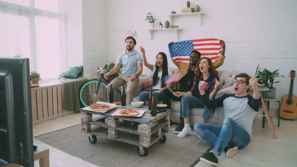 Group of diverse friends cheering for a sports event with American flags indoors.