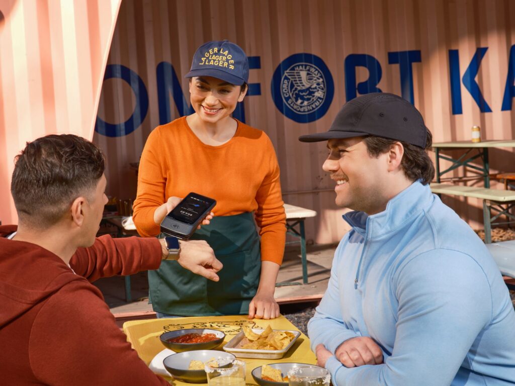 Friends using smartwatch for contactless payment at an outdoor cafe, assisted by a smiling waitress.