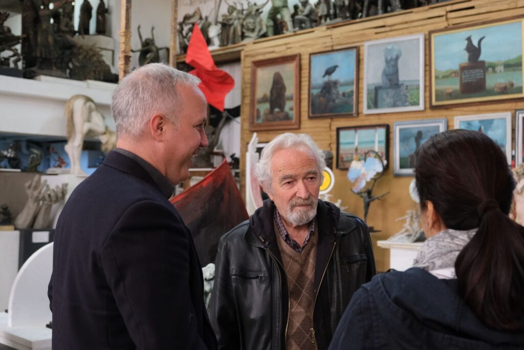 Three adults conversing in a sculpture workshop, surrounded by art pieces and paintings.