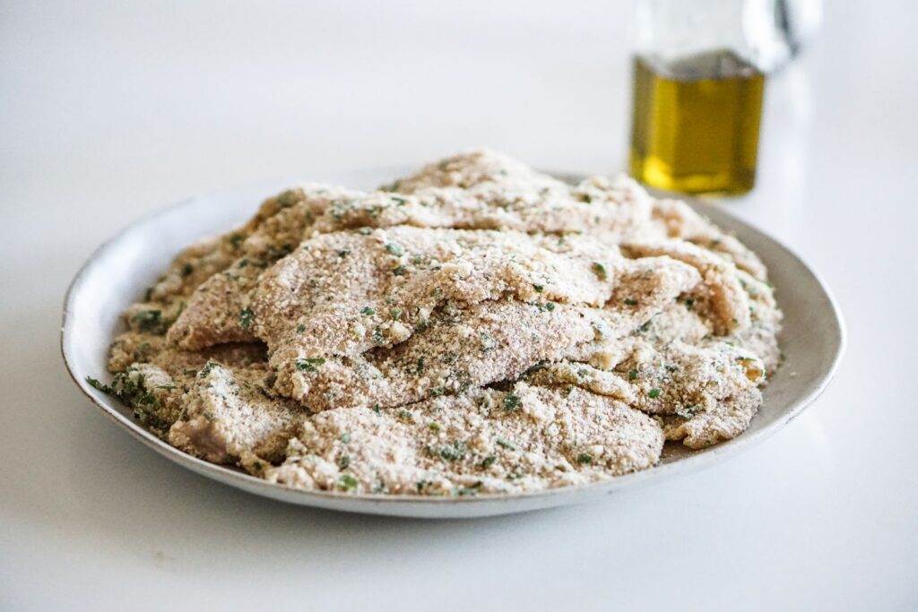 Plate of breaded chicken schnitzel, ready for cooking, with olive oil bottle in background.