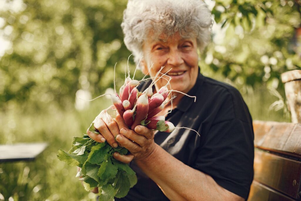 Senior woman smiling while holding a bunch of freshly harvested radishes outdoors in a garden.