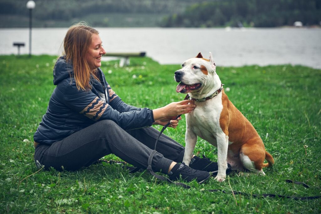 A woman sits on the grass with her American Staffordshire Terrier by a lake, happy and relaxed.