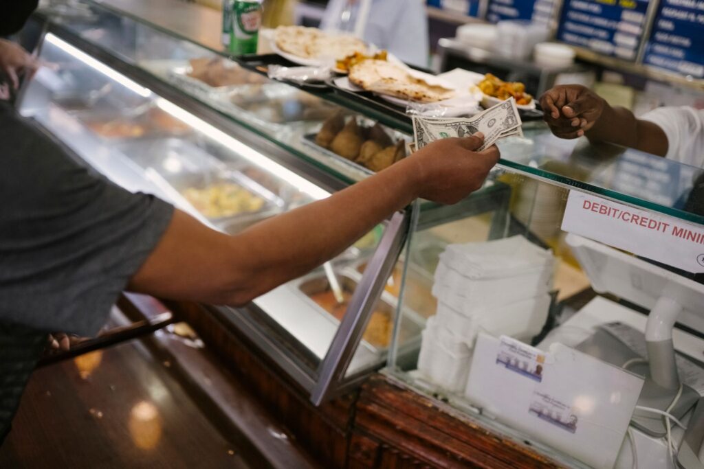 Crop faceless person standing near checkout buying food and passes money for dinner in cafe