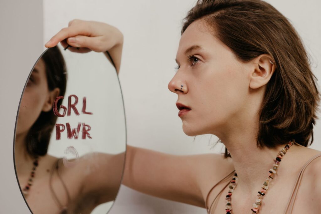 A young woman gazes into a mirror with 'GRL PWR' written on it, emphasizing empowerment.