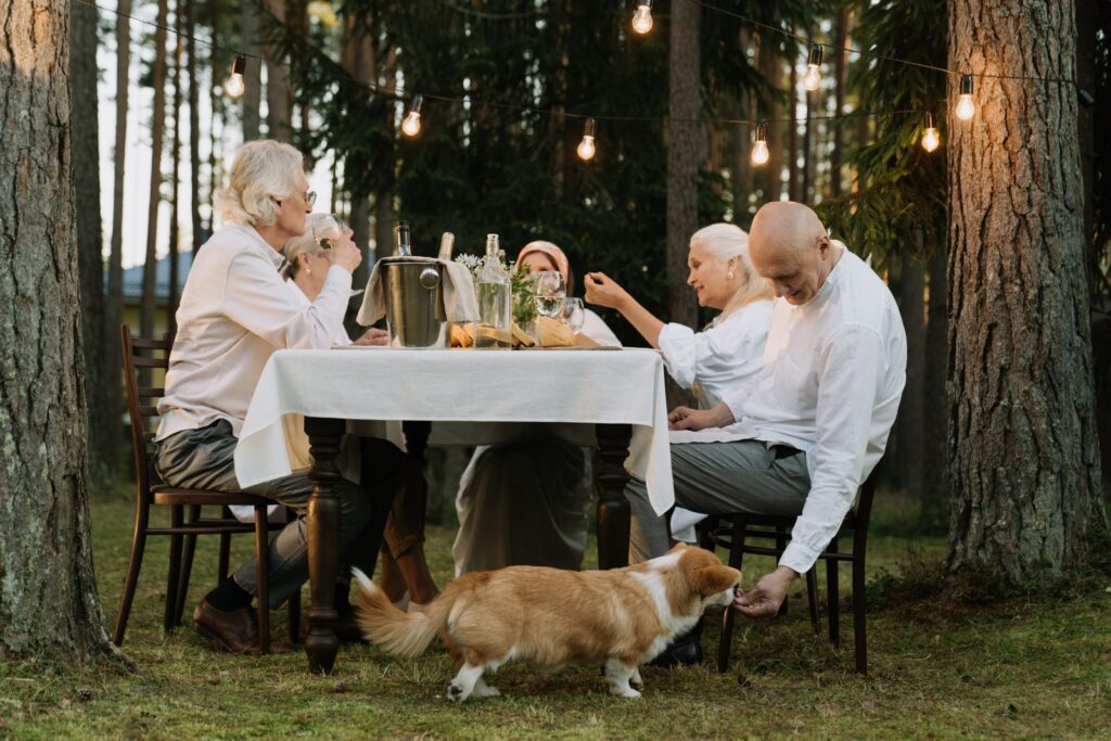 Seniors enjoying an outdoor dinner with a corgi under string lights in a forest setting.