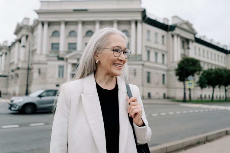 A senior woman in a white blazer smiling while walking outdoors near a historic building.