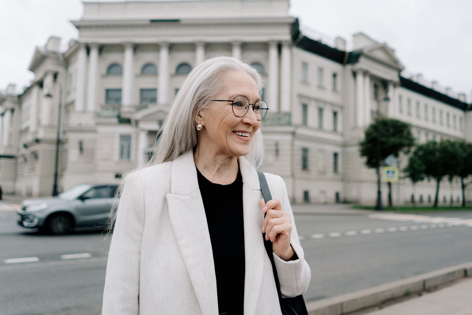 A senior woman in a white blazer smiling while walking outdoors near a historic building.