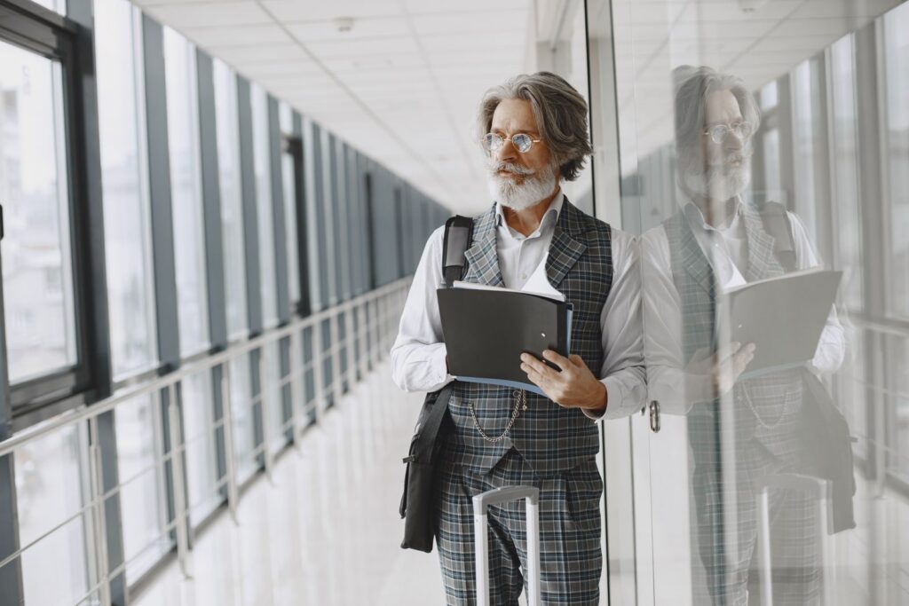 Elderly bearded man in a gray suit holding a folder in a modern glass hallway.