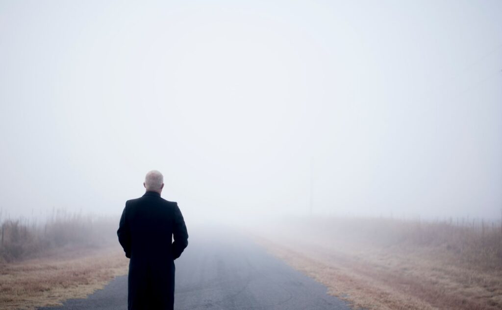 A lone man in a black coat stands on a foggy road, embodying solitude and introspection.