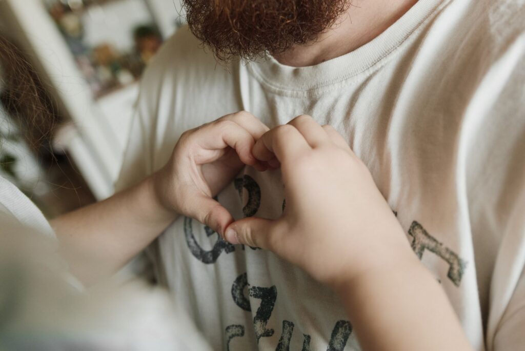 A child making a heart shape with hands on a bearded father's chest, expressing love and affection.