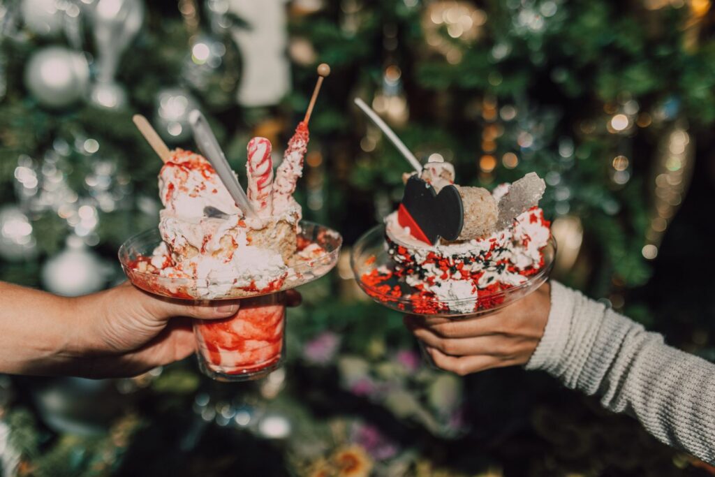 Two hands holding festive ice cream desserts with a Christmas theme, featuring colorful sprinkles and props.