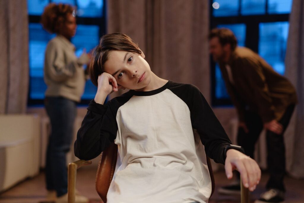 A young boy in a black and white shirt looks sad as his parents argue in the background.