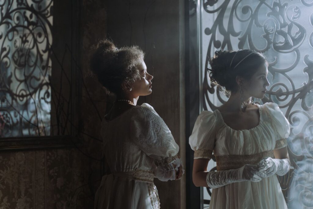 Two women in elegant historical dresses standing by an ornate window indoors.