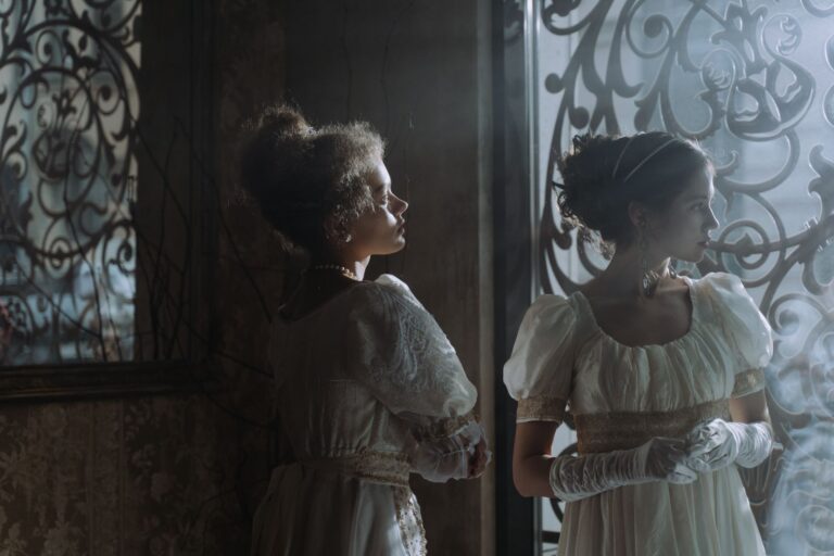 Two women in elegant historical dresses standing by an ornate window indoors.