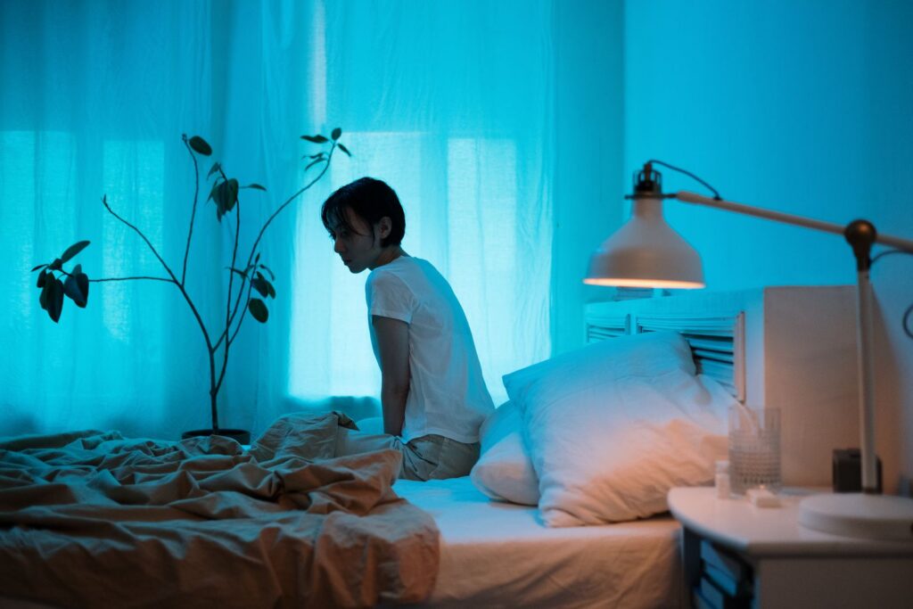 Contemplative woman sits on a bed in a blue-lit bedroom, reflecting solitude.