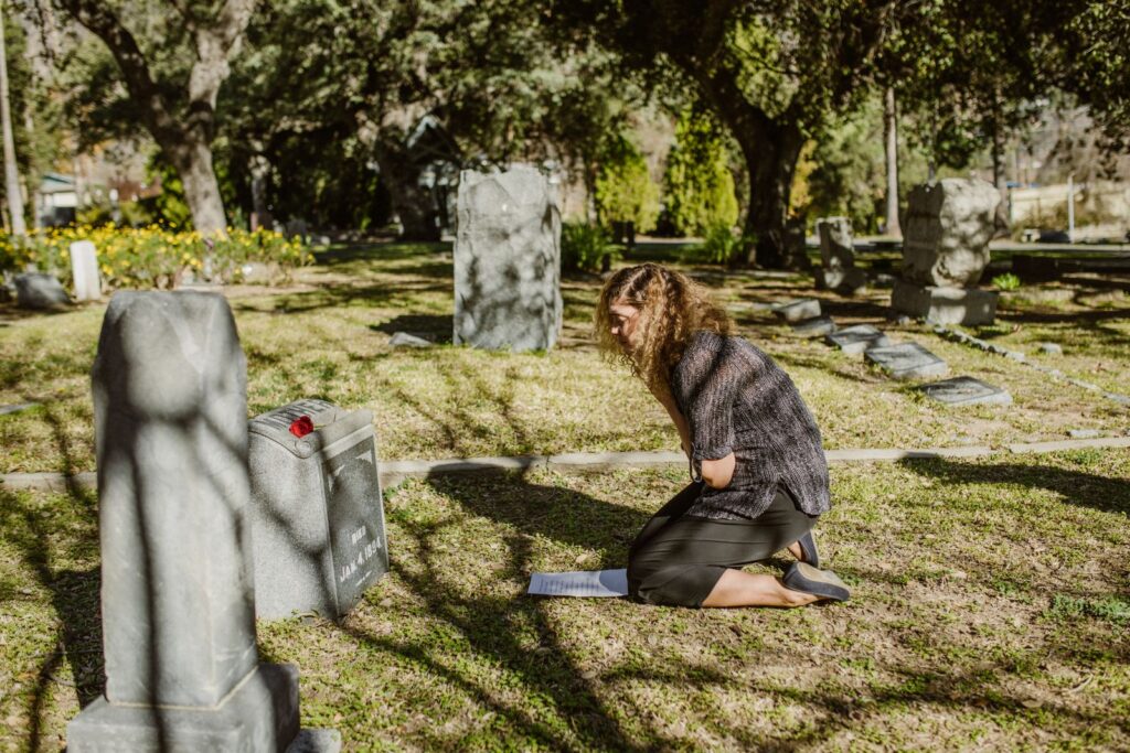 A woman kneels by a gravestone, expressing grief and loss in a sunlit cemetery.