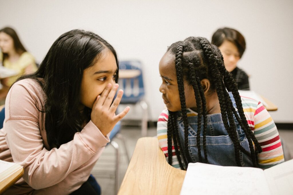 Two young girls whispering in a classroom setting, sharing a secret.
