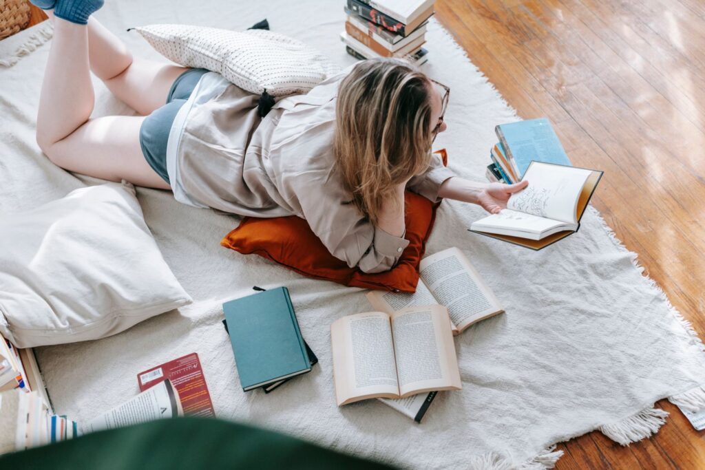 From above of young female in eyeglasses reading book while lying on plaid on floor