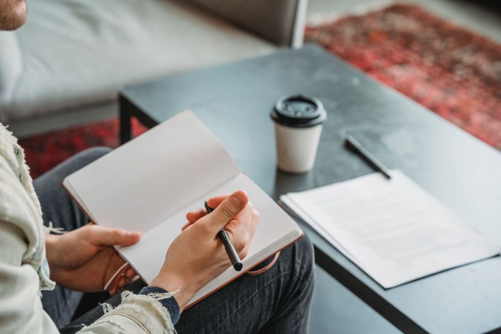 Close-up of a person writing in a notebook, with coffee on a table, indoors.