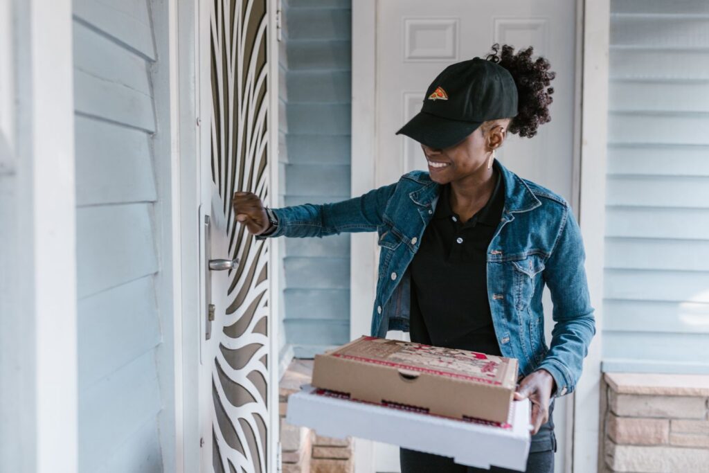 Smiling delivery woman knocking on front door holding pizza boxes.