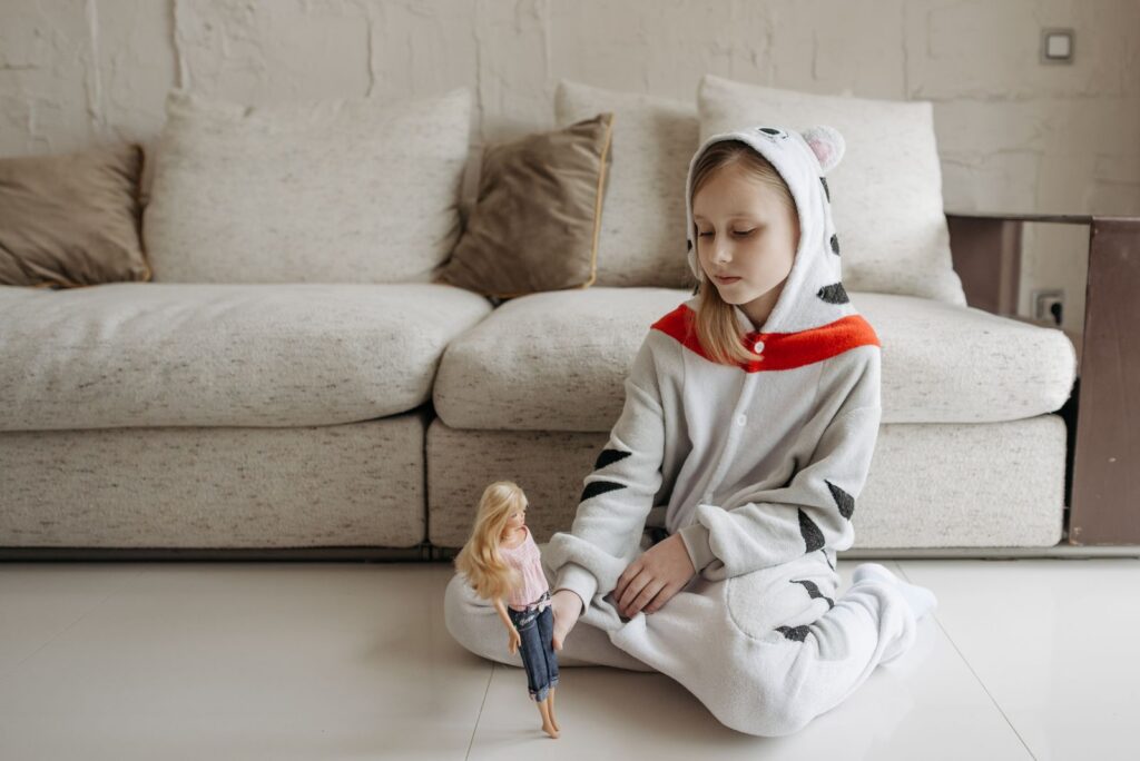 A young girl in a cozy onesie sits indoors playing with a doll near a comfortable couch.