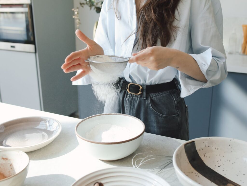 A close-up of a woman sifting flour into a bowl in a well-lit modern kitchen.