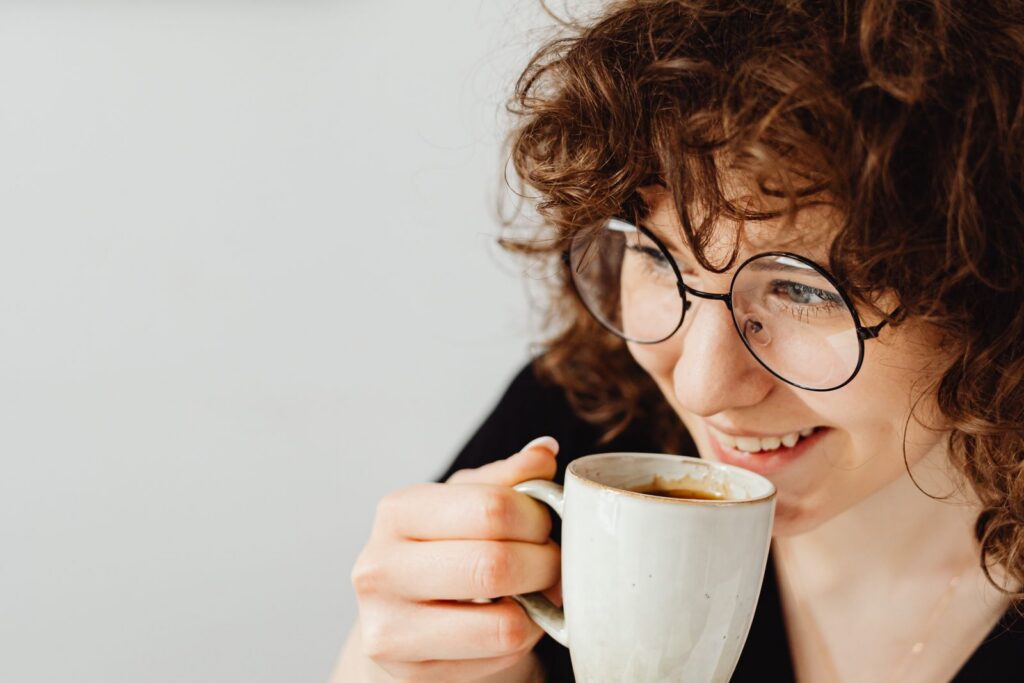 Smiling woman with curly hair and glasses savoring coffee in a cozy indoor setting.